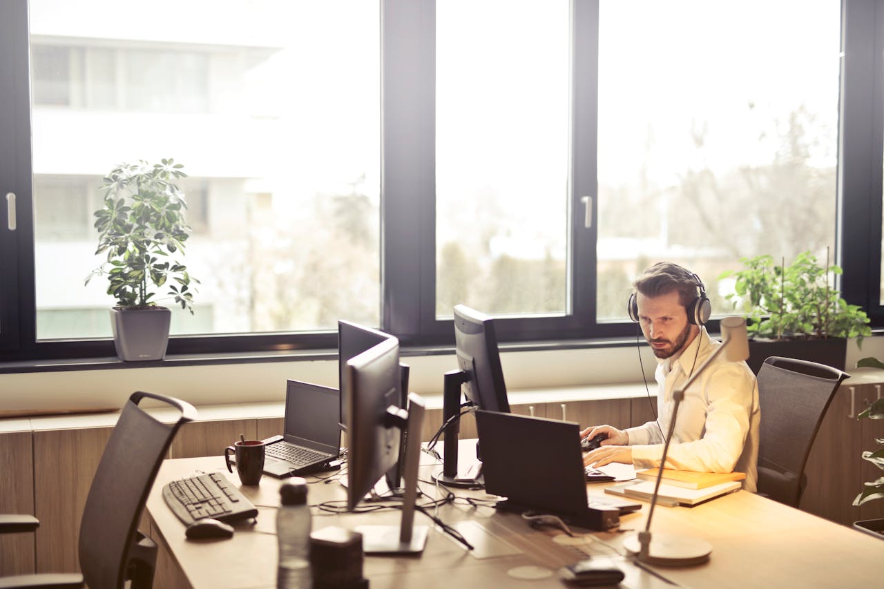 about-us A businessman sits at a desk using multiple computers and a headset in a well-lit modern office.